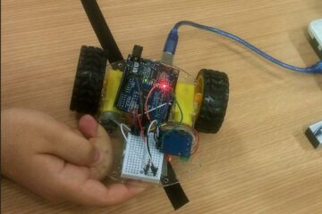 Homem segurando um robozino que parece com um carrinho feito de arduino e rodinhas.